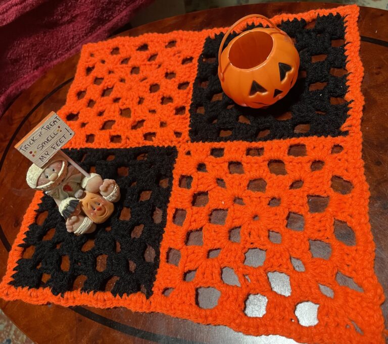 an orange and black checkerboard placement made out of crochet granny squares. On the placemat is a small jack-o-lantern bucket and a ceramic child mummy holding a sign that says "Trick or Treat Smell my Feet!"