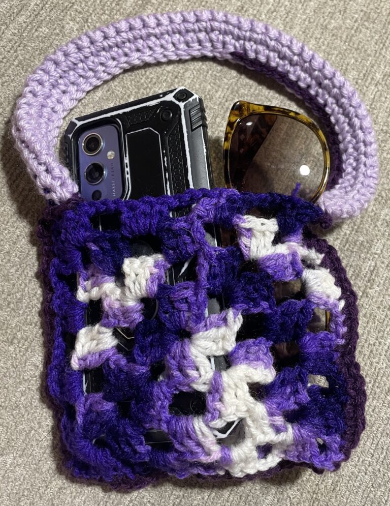 A small pocket bag made out of crochet granny squares with a light purple handle. The bag is holding a pair of sunglasses and a phone with a case on it.