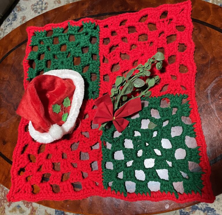 A red and green chckerboard placemat made out of four crochet granny squares. On the placemat there is a small Santa hat and a sprig of faux mistletoe